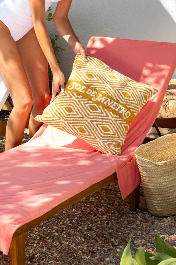 Woman placing a mustard and white diamond-pattern pillow on a wooden lounge chair draped in a coral Turkish towel, beside a straw basket