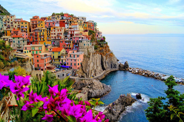 Pink, yellow and orange houses stacked on a cliff above a blue Mediterranean cove, with magenta bougainvillea in the foreground
