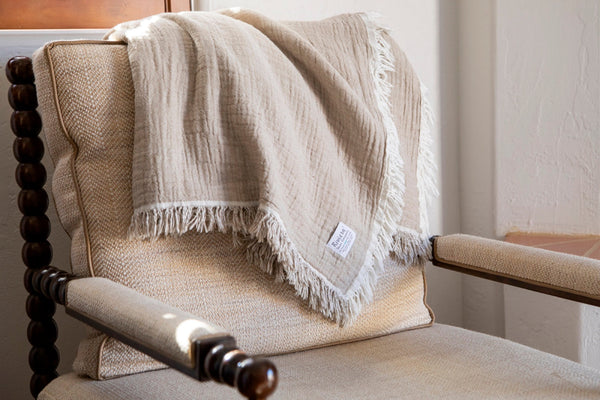 Beige woven throw blanket with fringed edges draped over a cream upholstered armchair