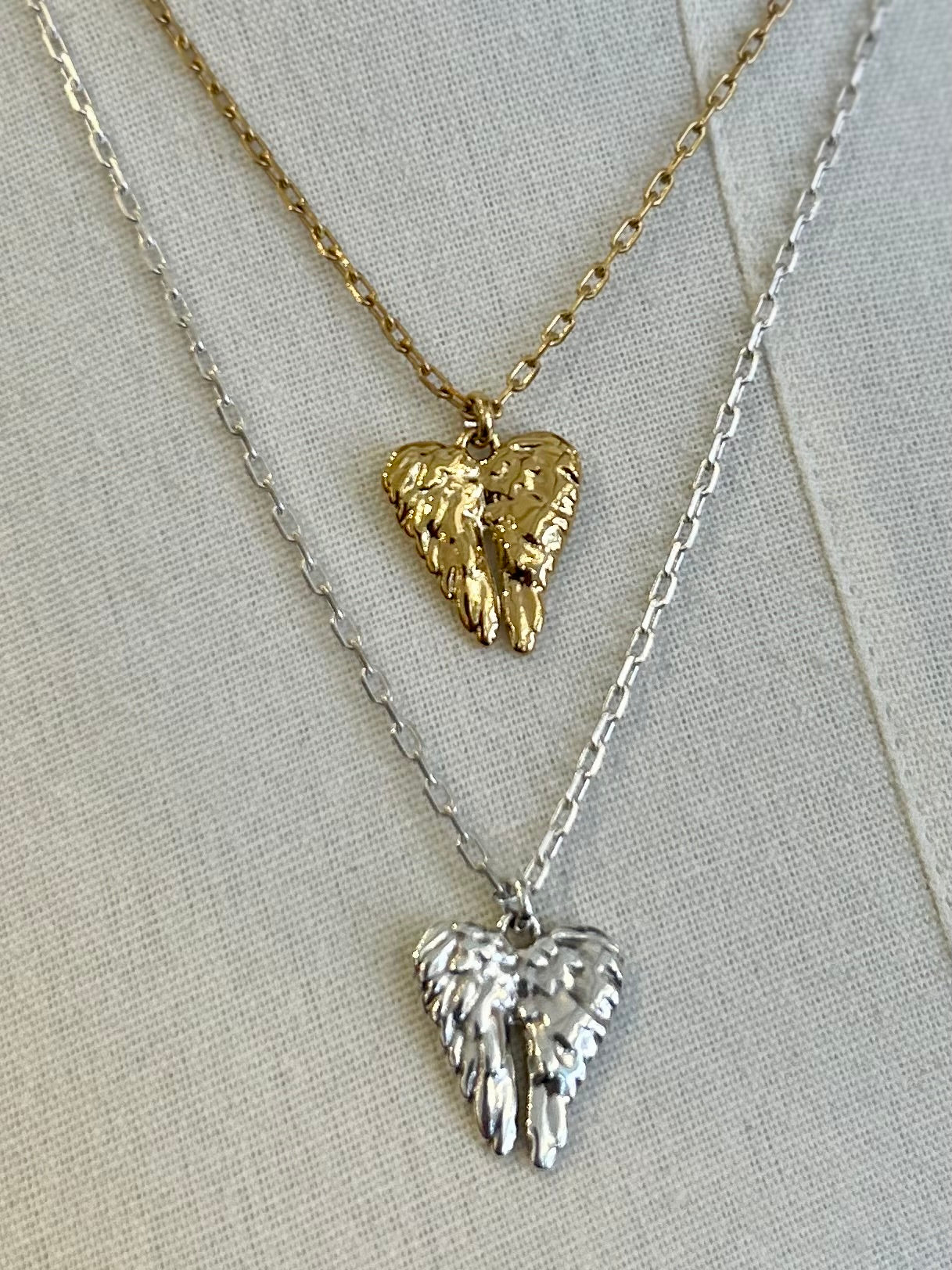 Two angel wing pendants on gold and silver necklaces against a light background