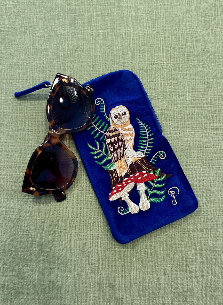 Embroidered owl design on a blue pouch with sunglasses on a wooden surface, styled