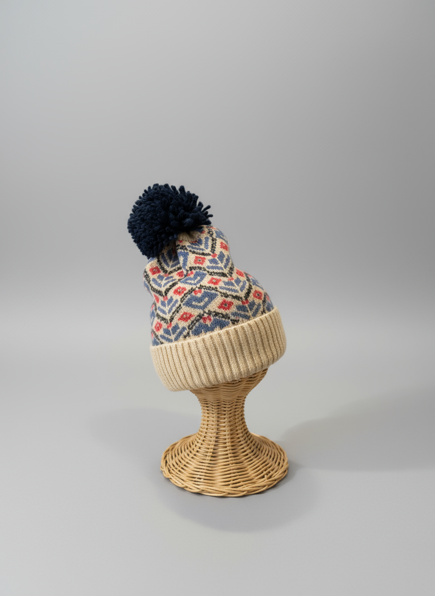 Knitted beanie with patterned top and pom-pom on a wicker stand against a gray background