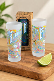 Two frosted glass tumblers with colorful illustrated coasters and the words 'Santa Barbara' and 'Barbaric' displayed in front of their branded cardboard packaging on a wooden surface.