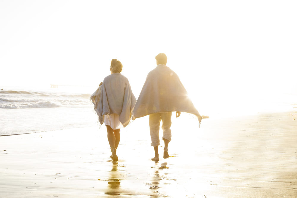 Couple walking barefoot along the shore at sunset wrapped in Whistler Turkish blankets
